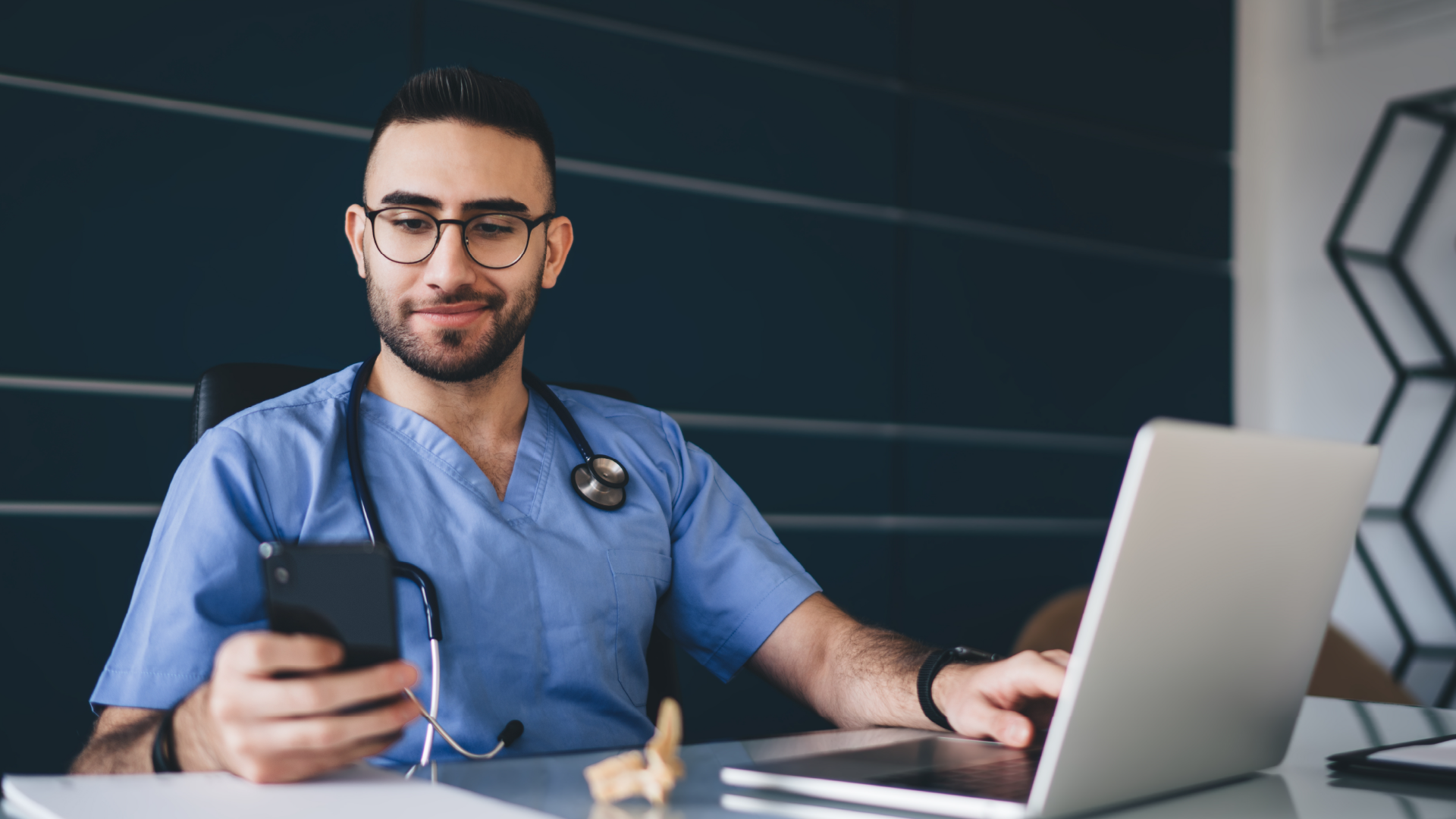A male doctor smiles as he looks at the smartphone he is holding in his right hand. They are sitting at a desk, with a laptop open, inside of a chic and modern looking doctors office.
